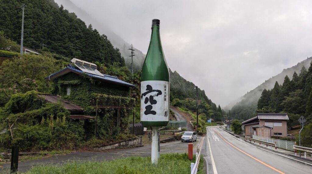 山間の道路沿いに大きな日本酒瓶の看板が立っている風景